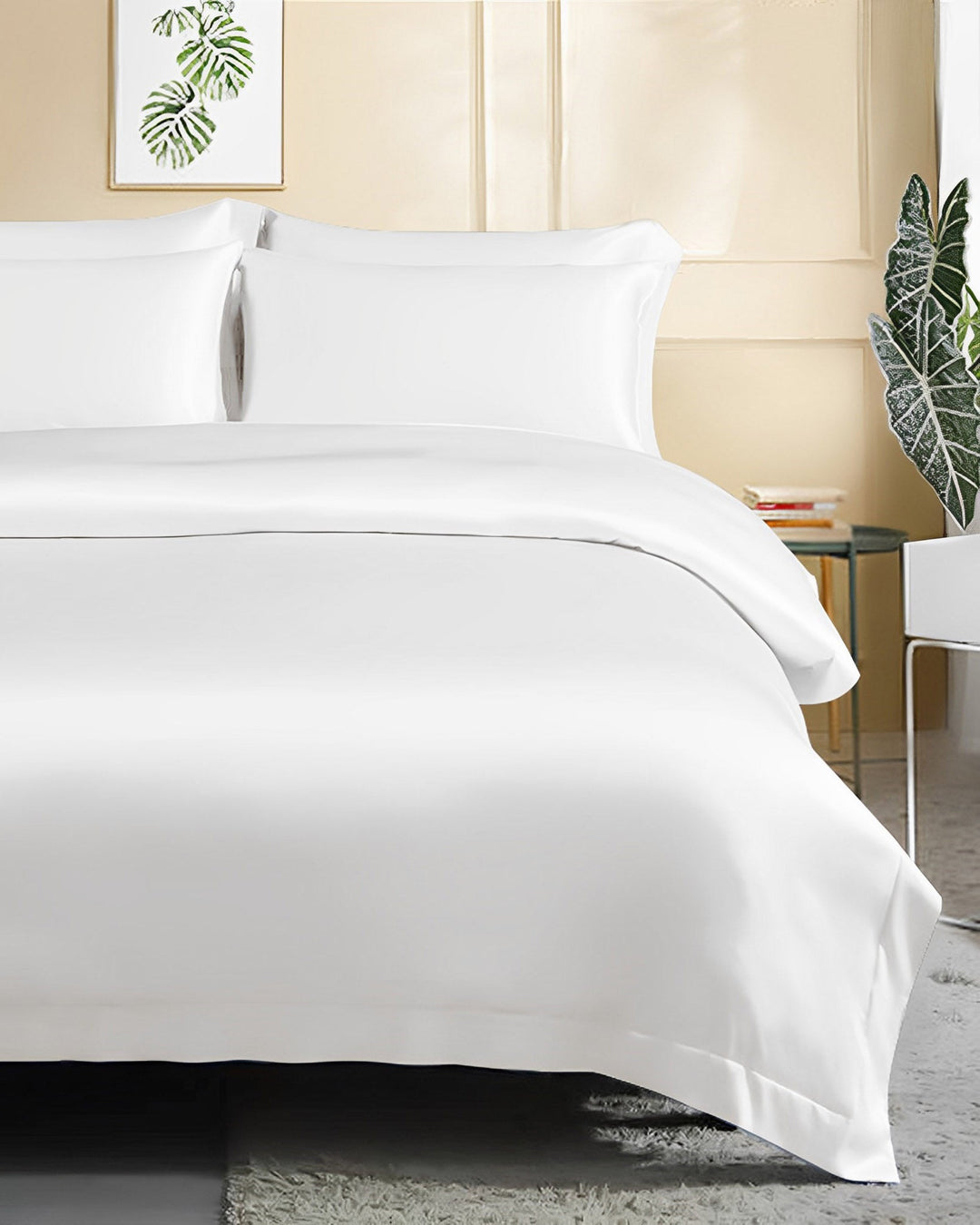 A neatly made bed featuring the SusanSilk 25 Momme Seamless Luxury Duvet Cover with Flange Border, complemented by white pillows and sheets, is set in a well-lit room with a beige wall adorned with a framed plant print, and green plants placed beside the bed.