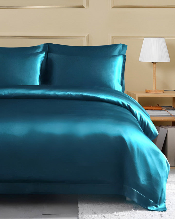 A neatly made bed with a turquoise SusanSilk 25 Momme Seamless Luxury Duvet Cover featuring a flange border and matching pillows, next to a light wooden nightstand holding a lamp with a white shade.