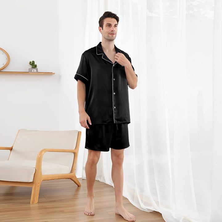 A person in SusanSilk's 25 Momme Short Silk Pajamas Set stands by a chair in a bright room with sheer white curtains.