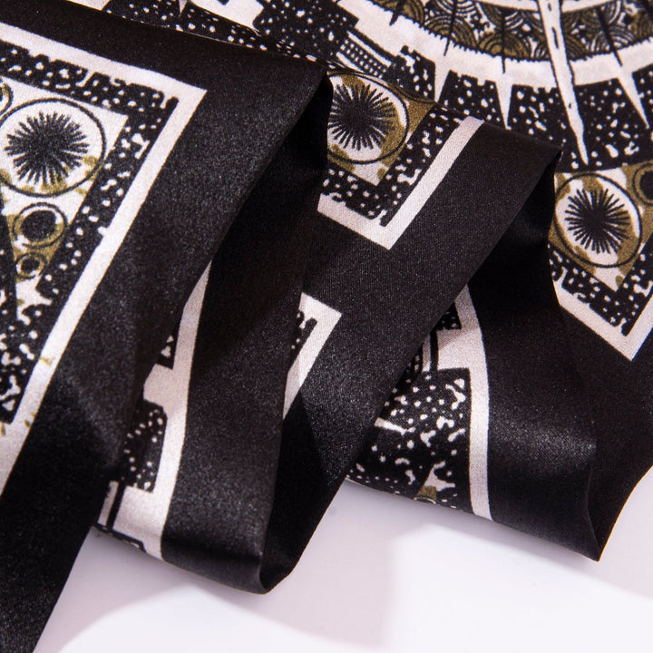 Close-up of a folded Black Compass Narrow Long Silk Scarf by SusanSilk featuring intricate black and white patterns, including circular and geometric shapes. The scarf appears silky and shiny.