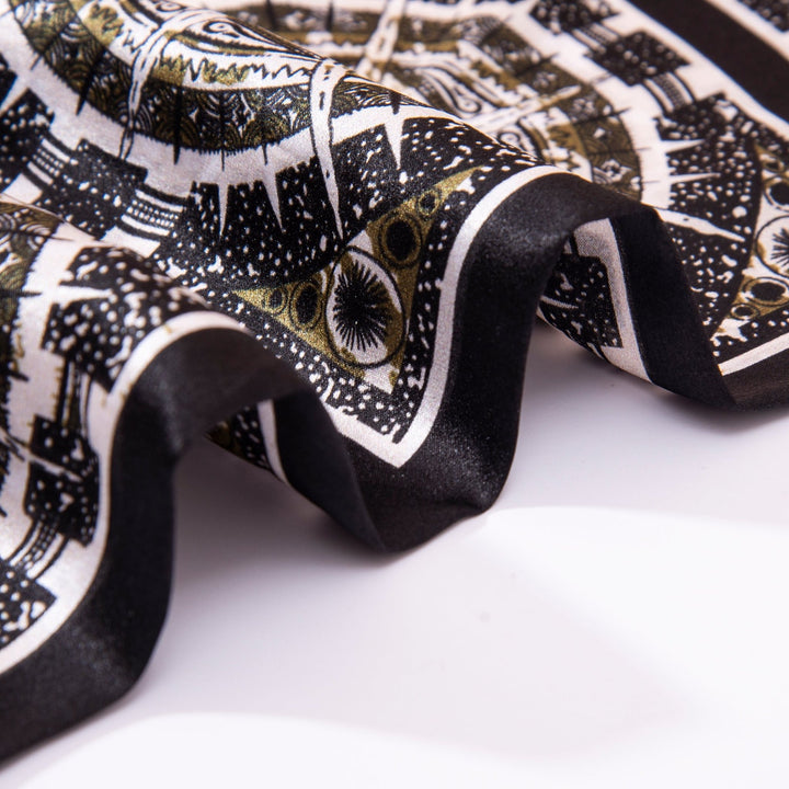 Close-up of the SusanSilk Black Compass Narrow Long Silk Scarf, showcasing its black and white pattern with gold accents, artfully folded and draped on a white surface.