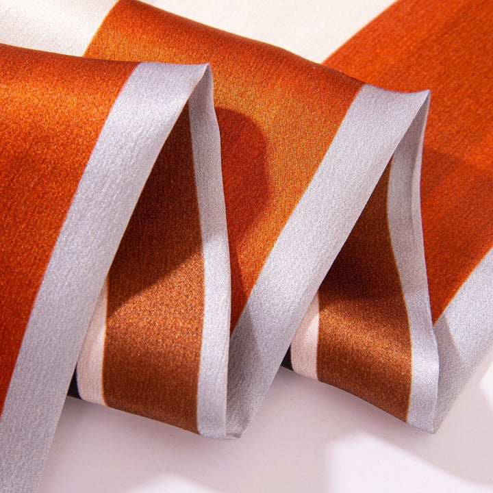 A close-up of the Color Strips Narrow Long Silk Scarf by SusanSilk, featuring folded silk fabric with alternating stripes of orange, white, and silver, showcasing its smooth texture and striking color contrast.