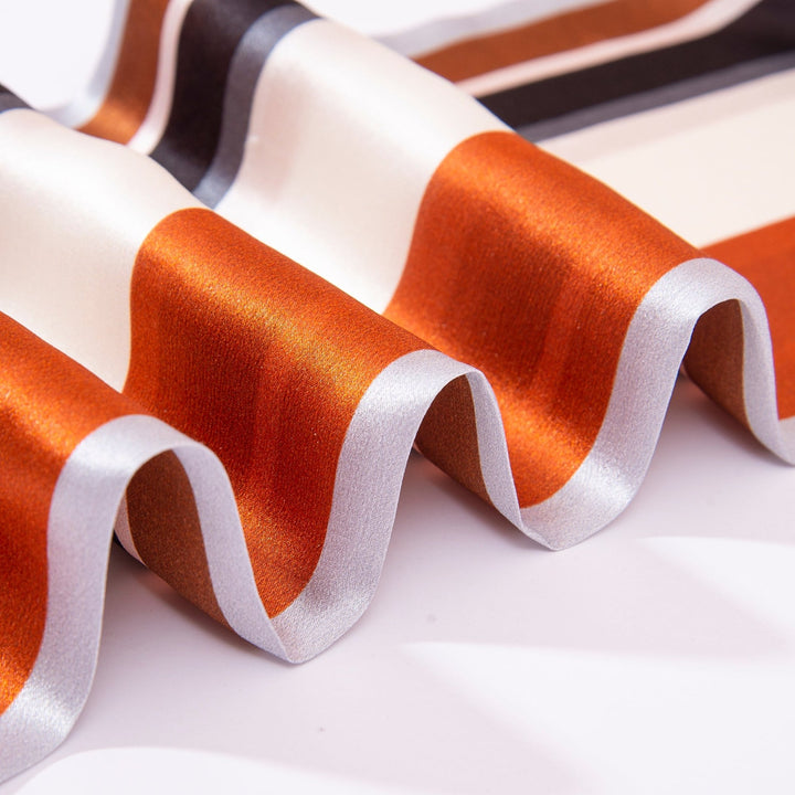 Close-up of a wavy, multi-colored fabric. The Color Strips Narrow Long Silk Scarf from SusanSilk features shiny surfaces with alternating stripes of orange, white, black, and light gray. This scarf beautifully shimmers in the light, highlighting its rich texture and vibrant hues.