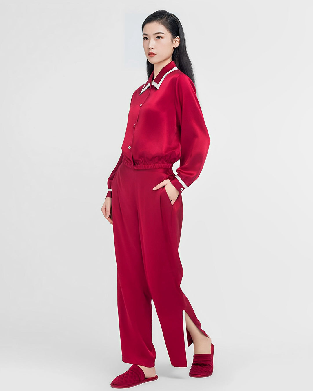 A woman stands against a plain background wearing the French Vintage Style Red Silk Pajamas Set from SusanSilk, complete with a matching red long-sleeve top and pants that feature side slits. She has her hands in her pockets and is wearing red slippers. This luxurious outfit, made from high-quality silk, exudes both elegance and comfort.