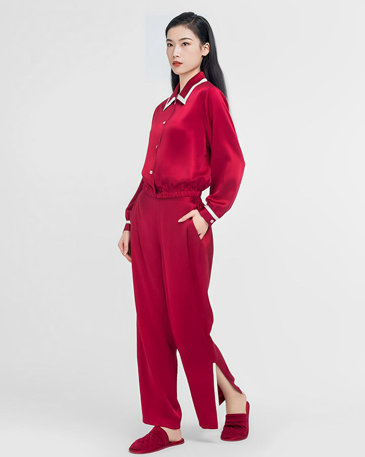 A woman stands against a plain background wearing the French Vintage Style Red Silk Pajamas Set from SusanSilk, complete with a matching red long-sleeve top and pants that feature side slits. She has her hands in her pockets and is wearing red slippers. This luxurious outfit, made from high-quality silk, exudes both elegance and comfort.