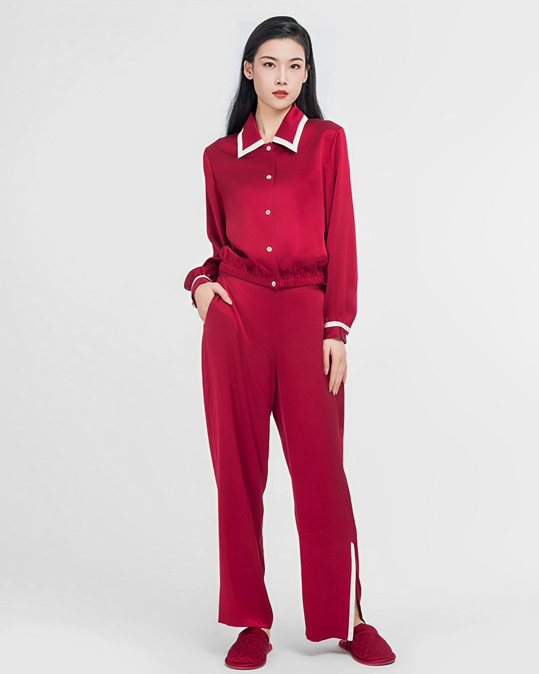 A person stands against a plain background wearing the SusanSilk French Vintage Style Red Silk Pajamas Set, which consists of a red silk shirt with white collar and cuffs, matching red pants with slits at the hems, and red slippers.