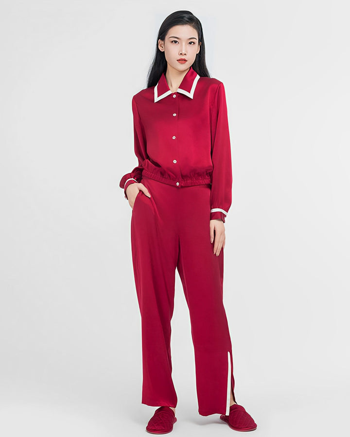 A person stands against a plain background wearing the SusanSilk French Vintage Style Red Silk Pajamas Set, which consists of a red silk shirt with white collar and cuffs, matching red pants with slits at the hems, and red slippers.