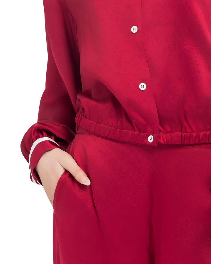 A person in the French Vintage Style Red Silk Pajamas Set by SusanSilk, characterized by a red silk long-sleeve shirt with white buttons and matching pants, is shown with their left hand in their pocket.