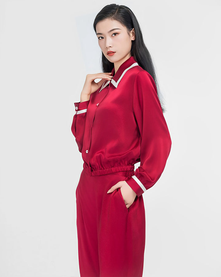 A woman with long black hair stands facing the camera, wearing a French Vintage Style Red Silk Pajamas Set from SusanSilk with white trim. She has one hand in her pocket and the other touching her collar. The silk fabric of her attire glistens under the light, against a plain white background.