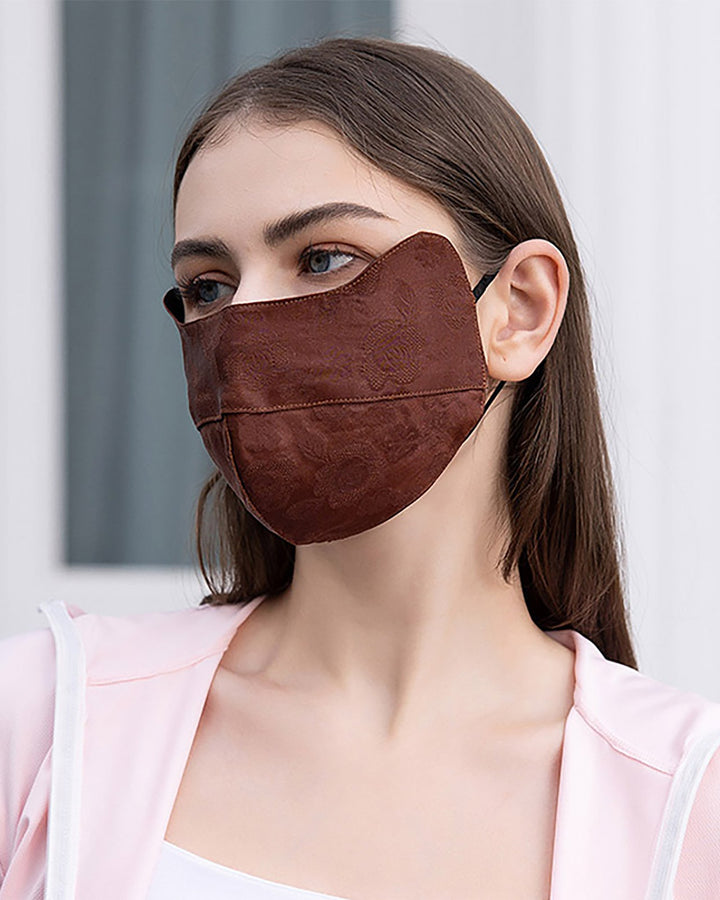 A woman with long brown hair is wearing a SusanSilk Jacquard Cloche Hat & Face Mask in brown silk and a light pink top. She is looking to the right, and the background is out of focus.
