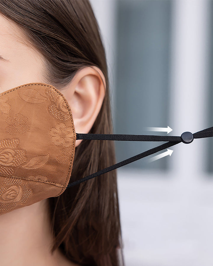 Close-up of a person wearing a floral-patterned face mask from the SusanSilk Jacquard Cloche Hat & Face Mask collection, featuring adjustable black ear loops. White arrows indicate the adjustment mechanism.