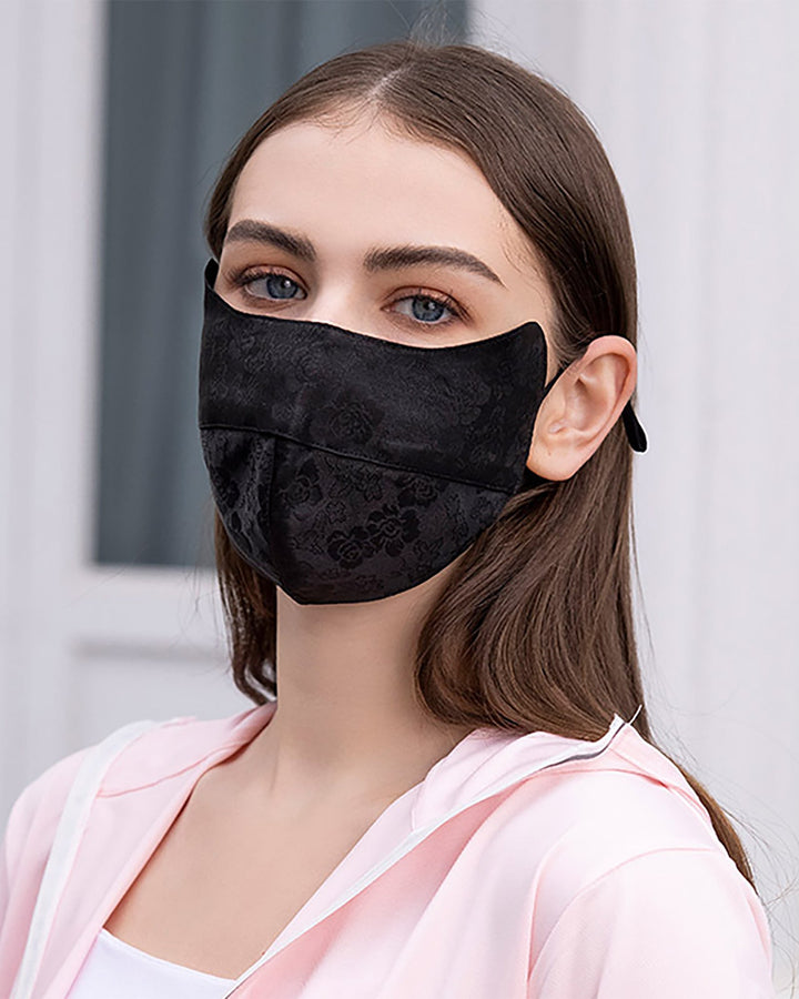 A woman with long brown hair, wearing a black face mask from SusanSilk's Jacquard Cloche Hat & Face Mask collection and a pink silk hoodie, stands in front of a light-colored background.