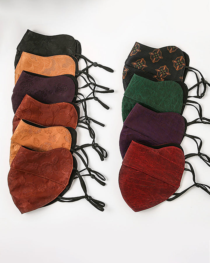 A collection of intricately patterned face masks, meticulously crafted from luxurious silk by SusanSilk, is displayed in the Jacquard Cloche Hat & Face Mask product range. These elegant masks come in an array of shades, including black, brown, orange, red, green, purple, and multicolor. They are arranged in two columns against a pristine white background.