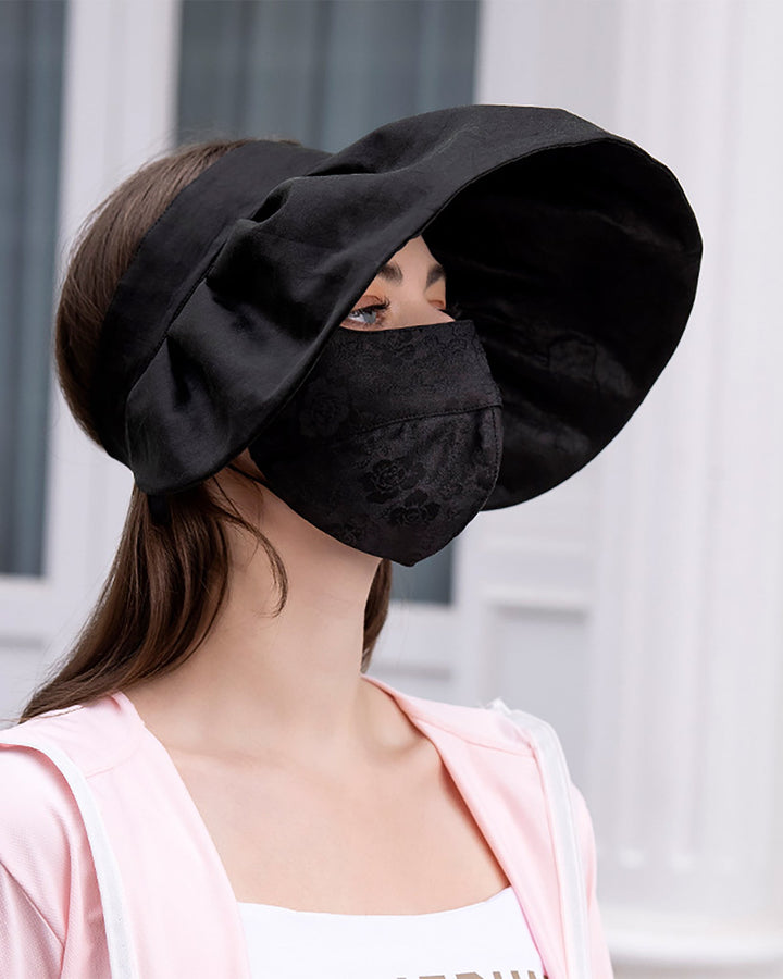 A person is wearing the Jacquard Large Brim Hat and matching face mask in black. They are dressed in a light pink silk jacket from SusanSilk.
