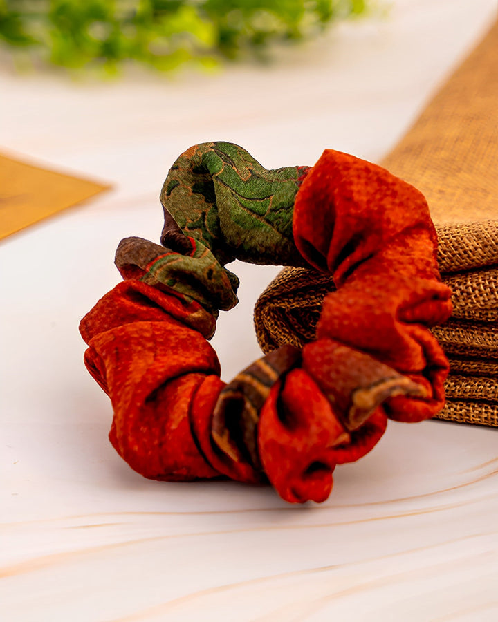 A luxurious Gambiered Canton Gauze Silk Scrunchie by SusanSilk, featuring a red and green patterned design, placed on a wooden surface next to a piece of burlap cloth.