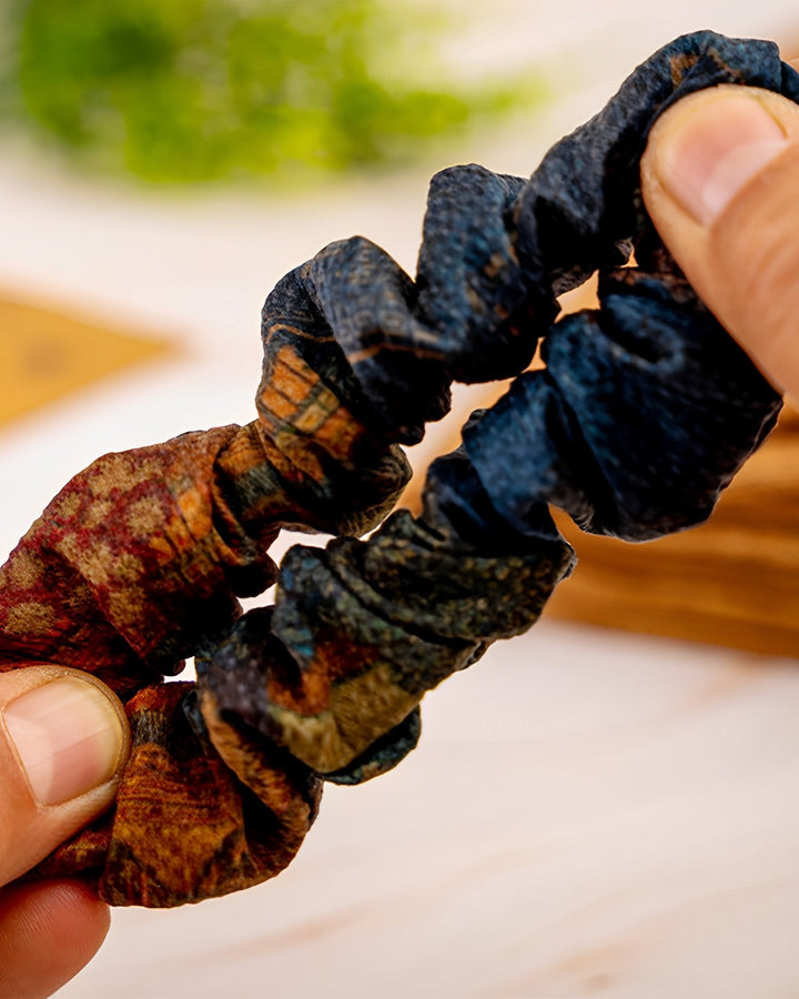 Close-up of two hands holding up two SusanSilk Gambiered Canton Gauze Silk Scrunchies (5cm), one fashioned from luxurious dark blue silk and the other adorned with multicolored brown and orange patterns. Blurred background.