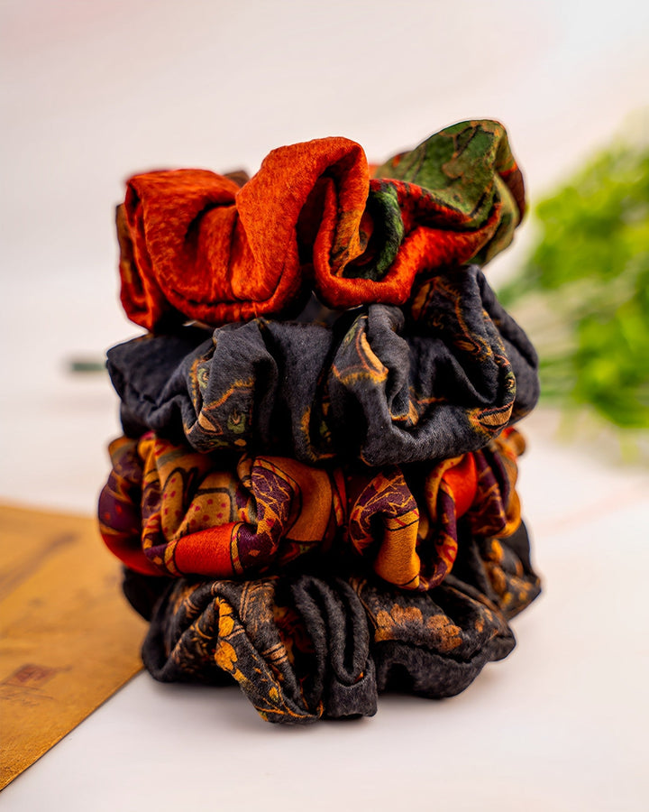 A vertical arrangement of Gambiered Canton Gauze Silk Scrunchies 5cm by SusanSilk in various colors and patterns, displayed on a surface with blurred greenery in the background.