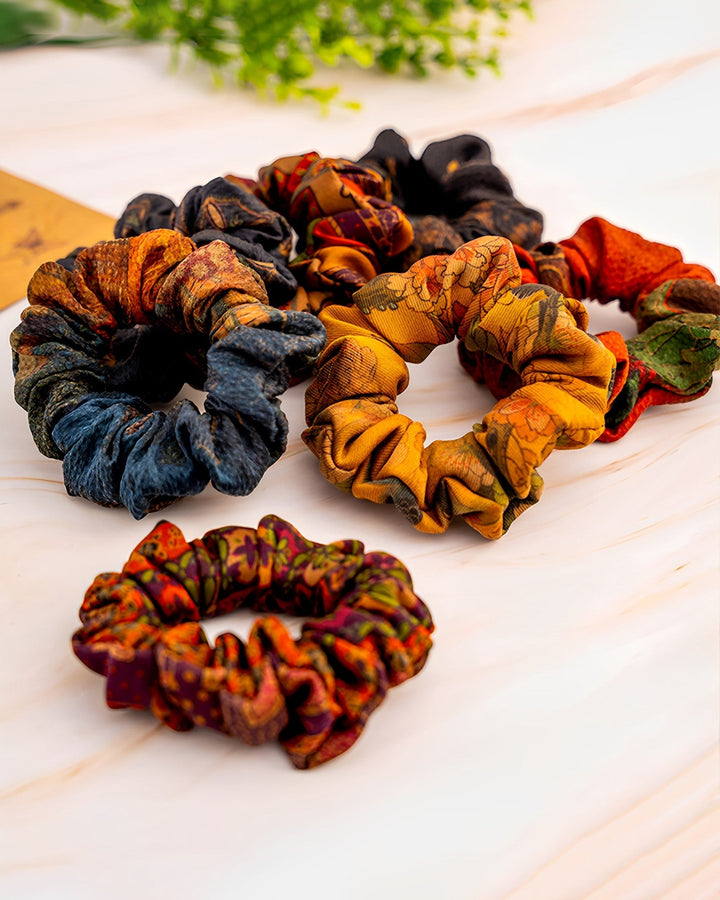 Five **SusanSilk Gambiered Canton Gauze Silk Scrunchies 5cm** in various colors and patterns are arranged on a light surface, with a green plant placed in the background, one scrunchie featuring luxurious silk.