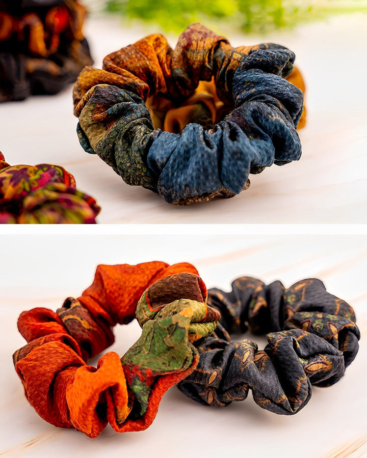 Two images of fabric scrunchies. The top image displays a single Gambiered Canton Gauze Silk Scrunchie (5cm) with a multicolored design from SusanSilk. The bottom image features two SusanSilk Gambiered Canton Gauze Silk Scrunchies (5cm)—one in orange with green and red accents, and the other in black adorned with intricate patterns.