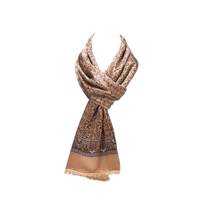 A light brown patterned silk scarf from SusanSilk, the Paisley Long Silk Brushed Scarf - Gold, is draped and tied in a knot. It features an intricate design with a border and fringe detailing along the lower edge.