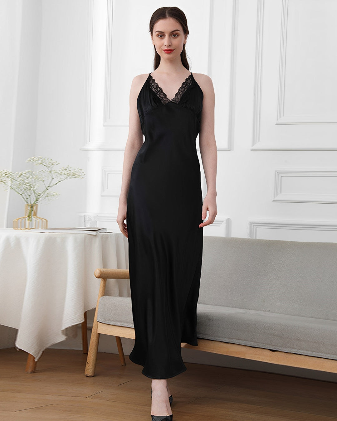 A woman is standing indoors wearing a SusanSilk Lace Long Silk Nightdress, a sleeveless black satin piece adorned with intricate lace detailing. The elegant scene is complemented by a wooden bench and a table draped with a white tablecloth and decorated with flowers, creating a striking contrast to her stunning nightdress.