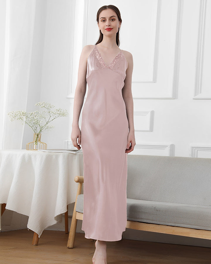 A woman stands indoors wearing a Lace Long Silk Nightdress from SusanSilk in light pink. She is smiling and has her arms by her sides. Behind her, there is a table with a white tablecloth and a vase with flowers, and a gray bench.
