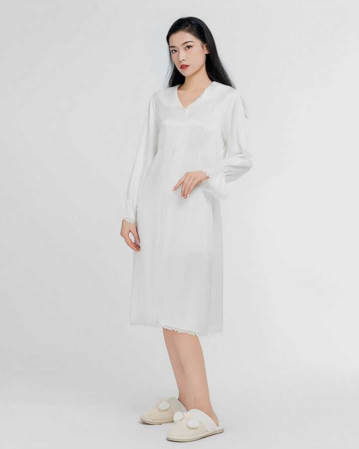 A woman is standing and wearing a Premium Lace Silk Nightdress by SusanSilk. She is also wearing white slippers with pom-poms. The background is plain and white.