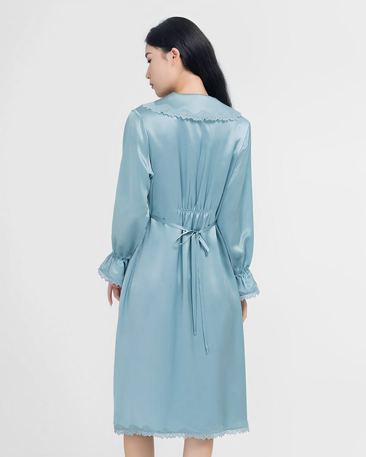 Woman in a Premium Lace Silk Nightdress by SusanSilk, seen from the back, against a plain white background. The silky blue knee-length dress has long sleeves, a sash tied at the waist, and scalloped lace edges.