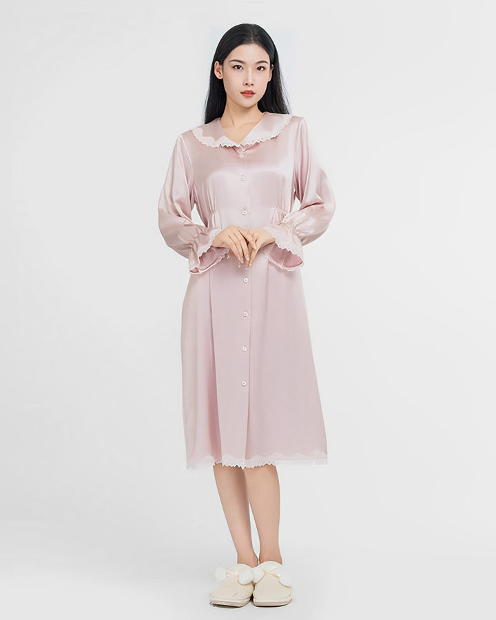 A woman stands wearing the SusanSilk Premium Lace Silk Nightdress, a light pink, long-sleeved silk garment adorned with white buttons and delicate lace trim. She pairs it with white slippers featuring bow accents against a plain white background.