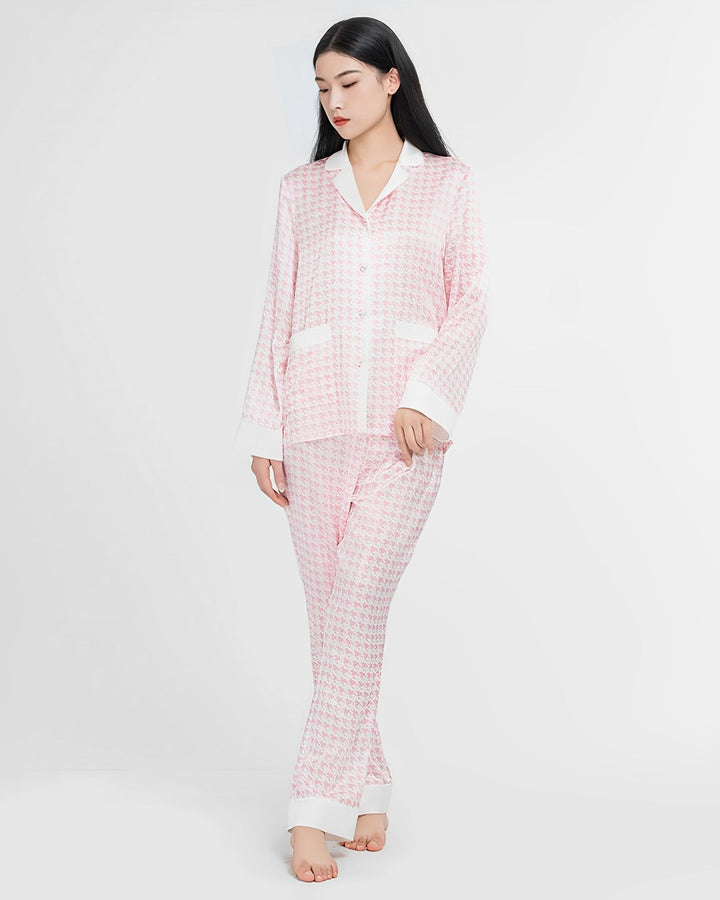 A person stands against a plain background wearing the Houndstooth Silk Pajamas Set from SusanSilk, featuring a collar, long sleeves, and full-length pants in pink and white.