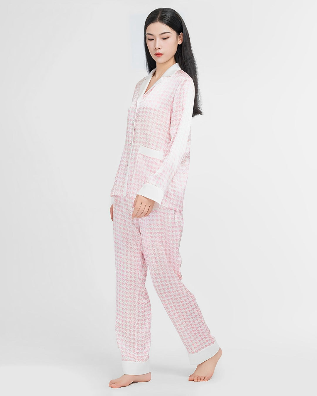 A woman with long black hair is wearing a light pink and white Houndstooth Silk Pajama set by SusanSilk. She is standing barefoot against a plain white background.