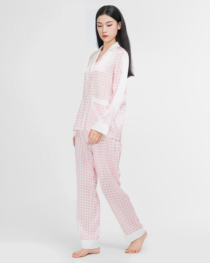 A woman with long black hair is wearing a light pink and white Houndstooth Silk Pajama set by SusanSilk. She is standing barefoot against a plain white background.