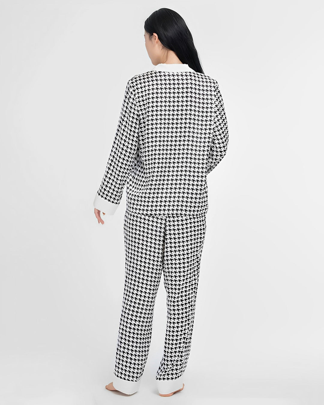 A person stands facing away from the camera, dressed in a matching SusanSilk Houndstooth Silk Pajamas Set, featuring a black-and-white houndstooth pattern and crafted from luxurious silk.
