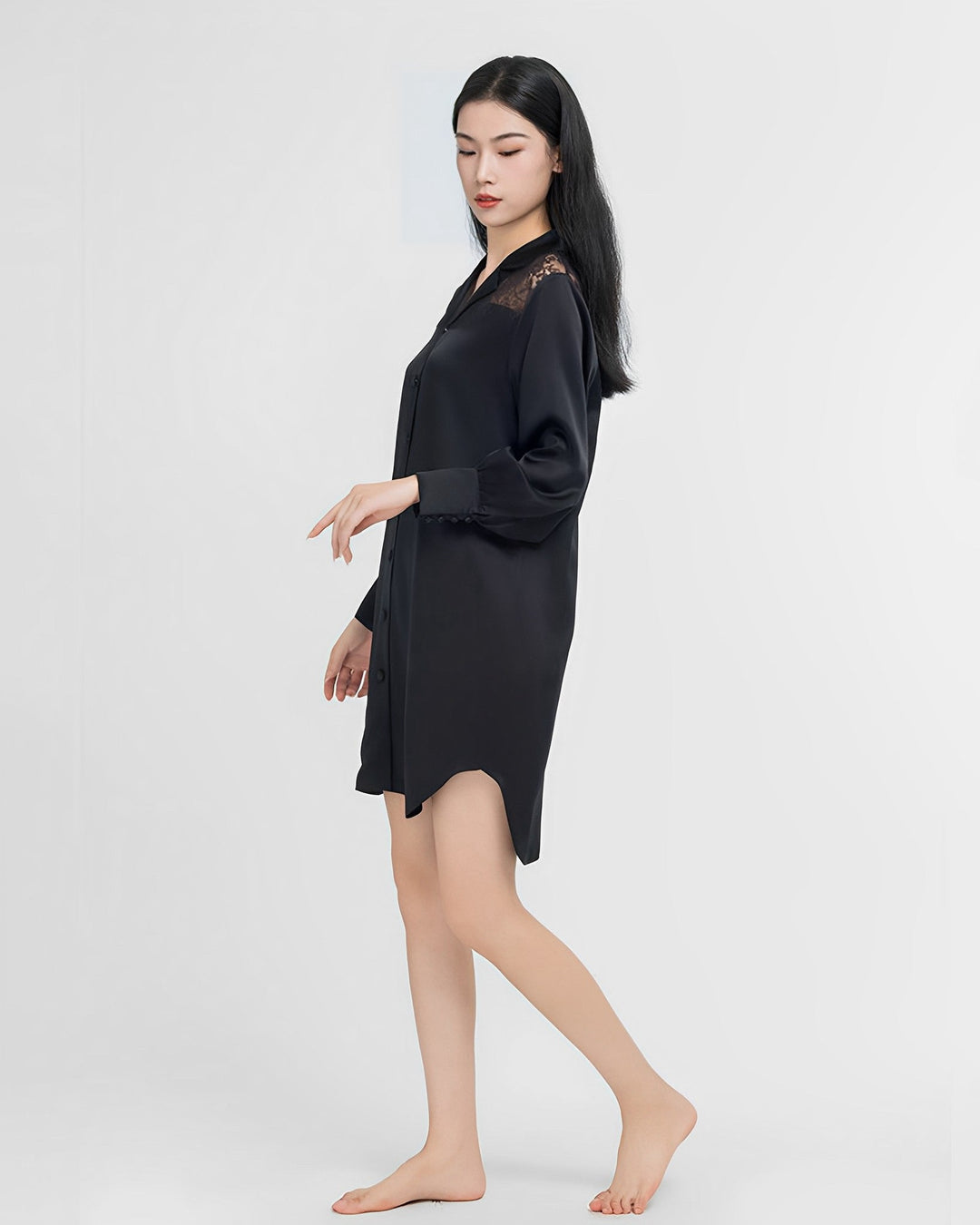 A woman in a sleek black Premium Lace Silk Shirtdress from SusanSilk, made of shimmering silk, stands barefoot against a plain white background, looking down and touching her fingertips.