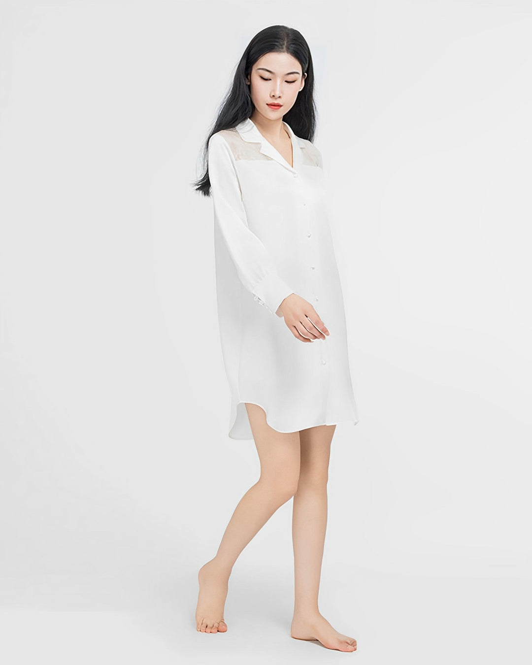 A person with long, straight dark hair is standing barefoot, wearing a white SusanSilk Premium Lace Silk Shirtdress against a plain white background.