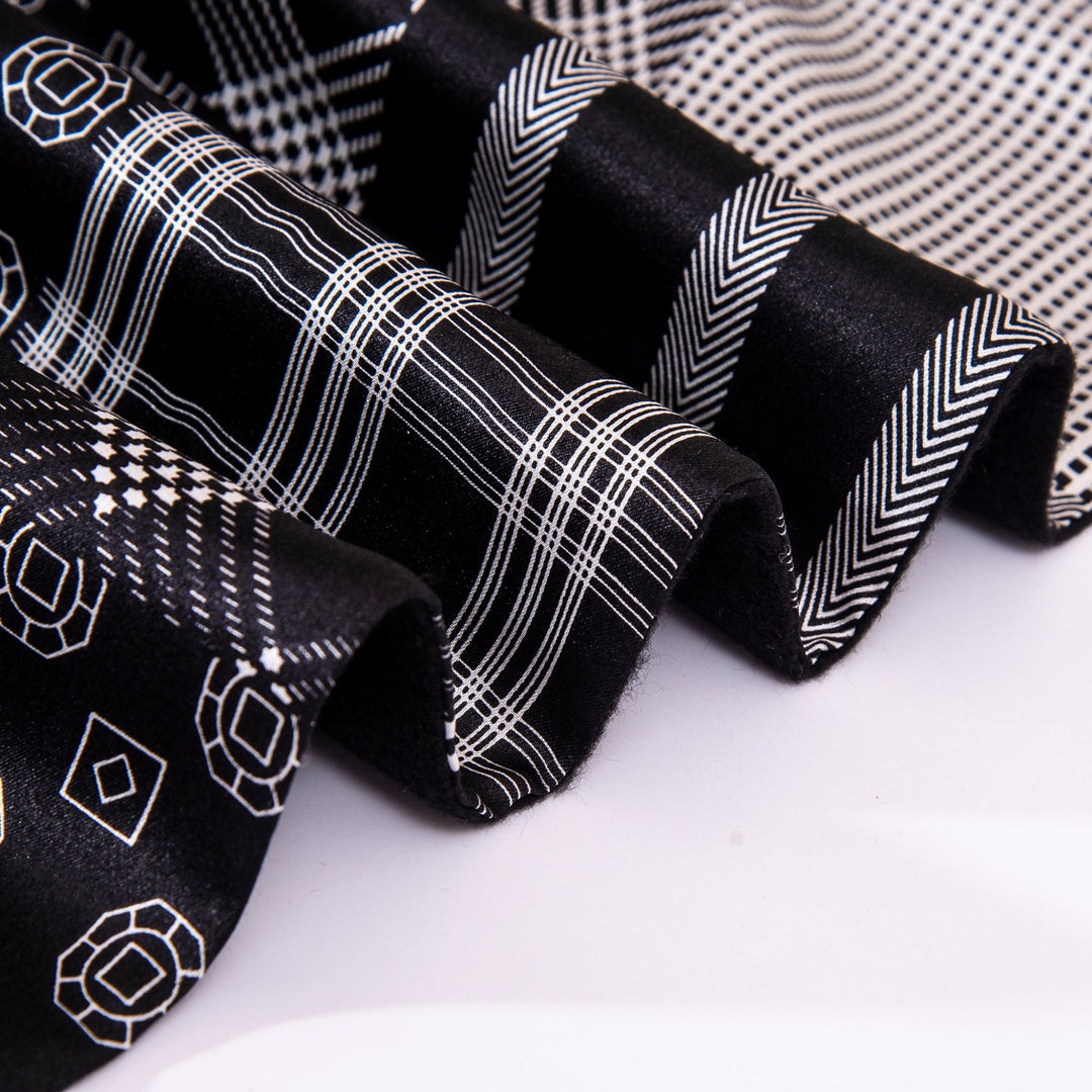 Close-up of SusanSilk's Mix Pattern Long Silk Brushed Scarf in black, featuring various white geometric patterns including stripes, hexagons, and chevrons. The luxurious silk fabric appears folded and layered.