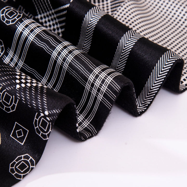 Close-up of SusanSilk's Mix Pattern Long Silk Brushed Scarf in black, featuring various white geometric patterns including stripes, hexagons, and chevrons. The luxurious silk fabric appears folded and layered.