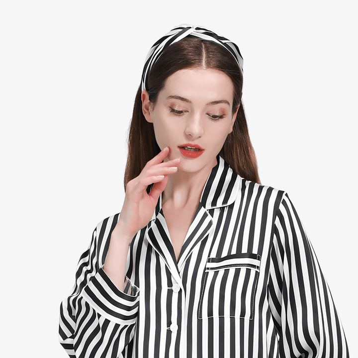 A woman with long brown hair wears a black and white striped silk pajama top and SusanSilk's Stripe Criss Cross Silk Headband, looking down with her hand gently touching her face.