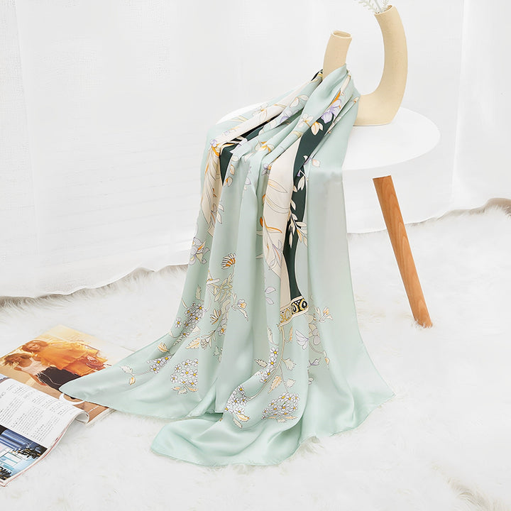 A SusanSilk Ornament & Floral Long Silk Crepe Scarf, in light green with floral patterns, gracefully draped over a white side table next to a magazine on the floor, all set atop a white fuzzy rug.