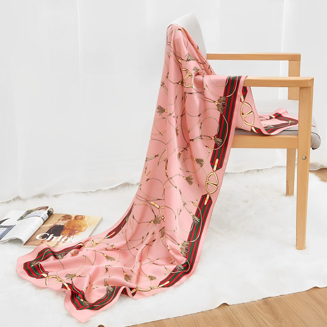 An Ornament & Floral Long Silk Crepe Scarf from SusanSilk, featuring a light pink hue and a chain pattern, is draped over a wooden chair. A magazine lies open on the floor beside the chair.