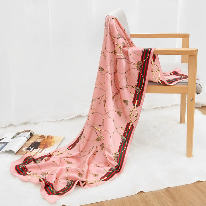 An Ornament & Floral Long Silk Crepe Scarf from SusanSilk, featuring a light pink hue and a chain pattern, is draped over a wooden chair. A magazine lies open on the floor beside the chair.
