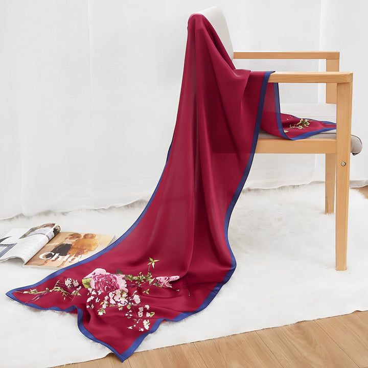 An Ornament & Floral Long Silk Crepe Scarf by SusanSilk, richly designed with red floral patterns, is draped over a wooden chair in a bright room featuring white curtains, a white rug, and magazines scattered on the floor.