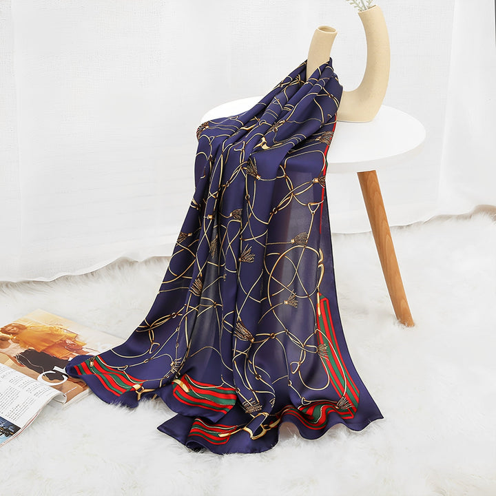 The Ornament & Floral Long Silk Crepe Scarf by SusanSilk, featuring a navy blue and gold pattern with red and green stripes, is draped over a white table adorned with a mannequin hand. A fashion magazine lies open on a fluffy white rug below.
