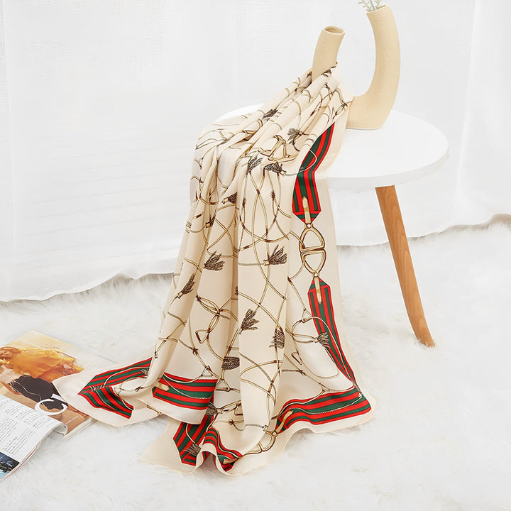 An Ornament & Floral Long Silk Crepe Scarf from SusanSilk, featuring beige tones with red, green, and black accents, is draped over a white table on a fluffy white rug next to an open magazine.