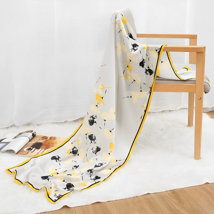 An Ornament & Floral Long Silk Crepe Scarf by SusanSilk, featuring a white and yellow floral pattern, is draped over a wooden chair with one end cascading onto a plush white rug on the floor. Nearby, there is an open magazine and another resting on the rug.