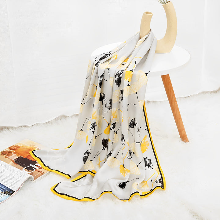 An Ornament & Floral Long Silk Crepe Scarf by SusanSilk, showcasing a lightweight yellow and black botanical pattern, is draped over a white side table on a white fur rug. A magazine and vase are also placed on the table.