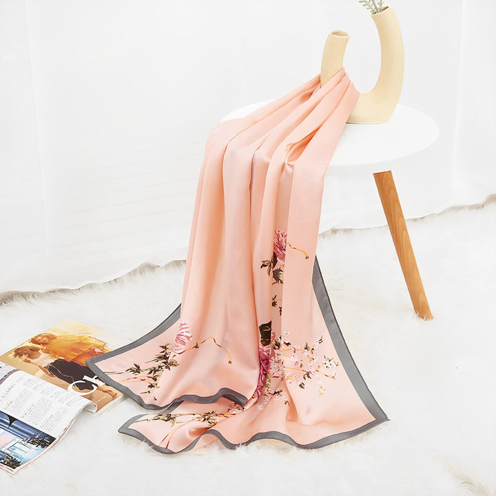 The Ornament & Floral Long Silk Crepe Scarf by SusanSilk, featuring peach-colored floral patterns and a gray border, is draped over a white table with wooden legs and sits next to an open magazine on a fluffy white rug.