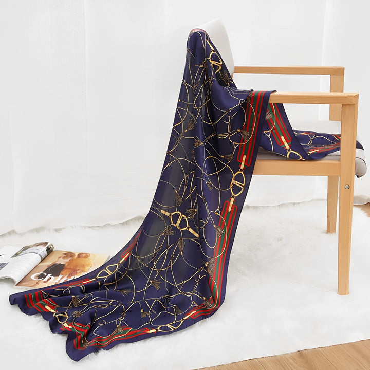 An Ornament & Floral Long Silk Crepe Scarf by SusanSilk, featuring a dark blue background with a gold chain pattern and red-green stripes, is draped over a wooden chair. A magazine is on the floor beside the chair, which sits on a white plush rug.