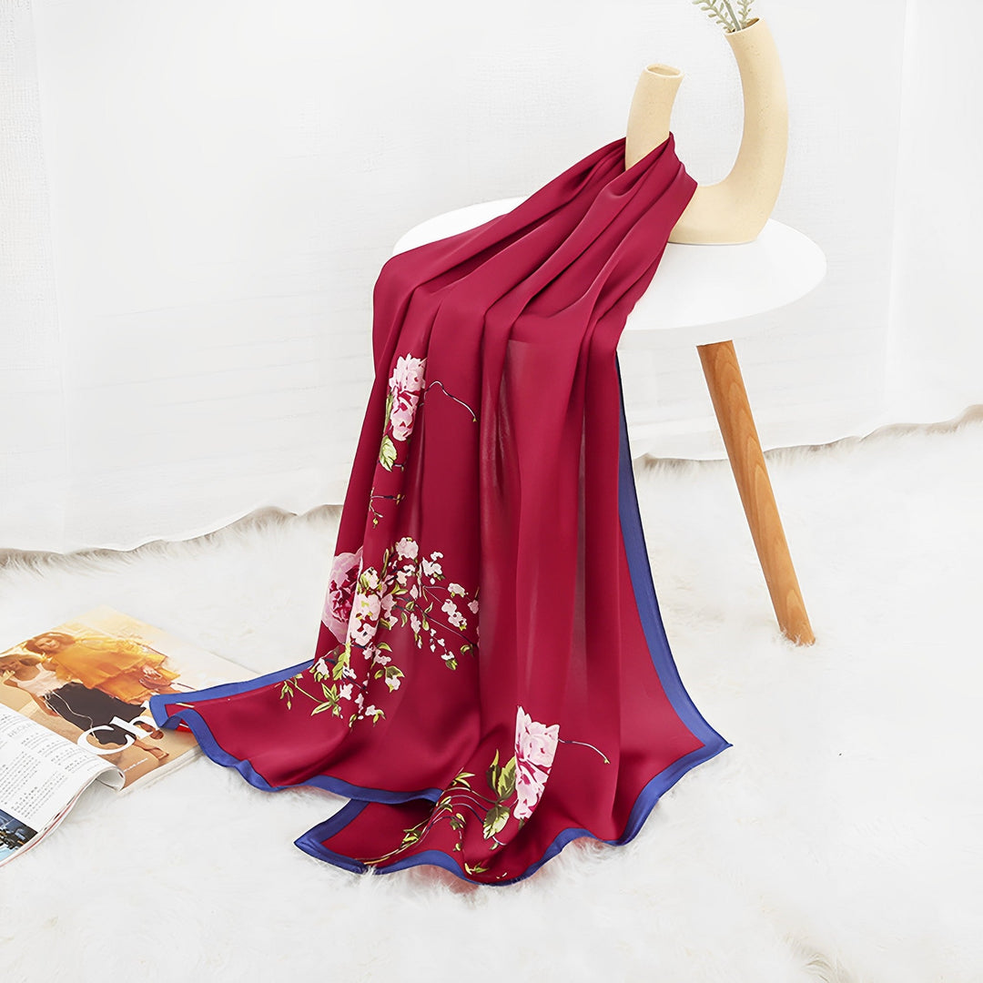 A SusanSilk Ornament & Floral Long Silk Crepe Scarf is draped over a white table with wooden legs. A rolled-up magazine lies on the white, fluffy rug beneath, adding to the luxurious ambiance.