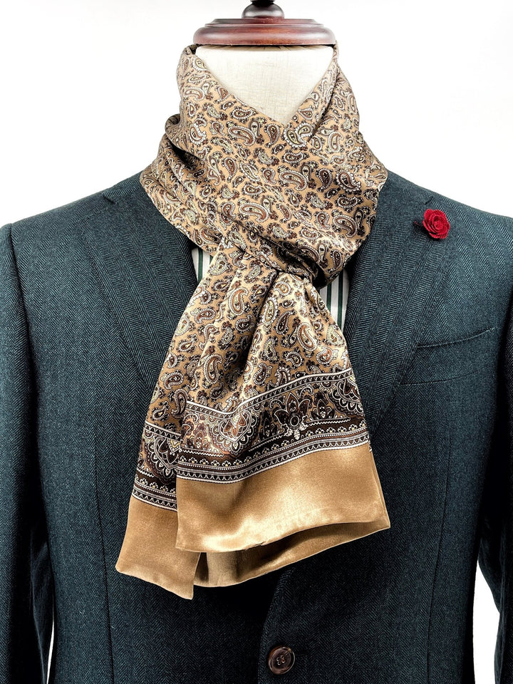 A mannequin dressed in a dark teal suit jacket adorned with a red lapel pin is wearing the Paisley Series Long Silk Scarf from SusanSilk, featuring a beige and brown paisley pattern.
